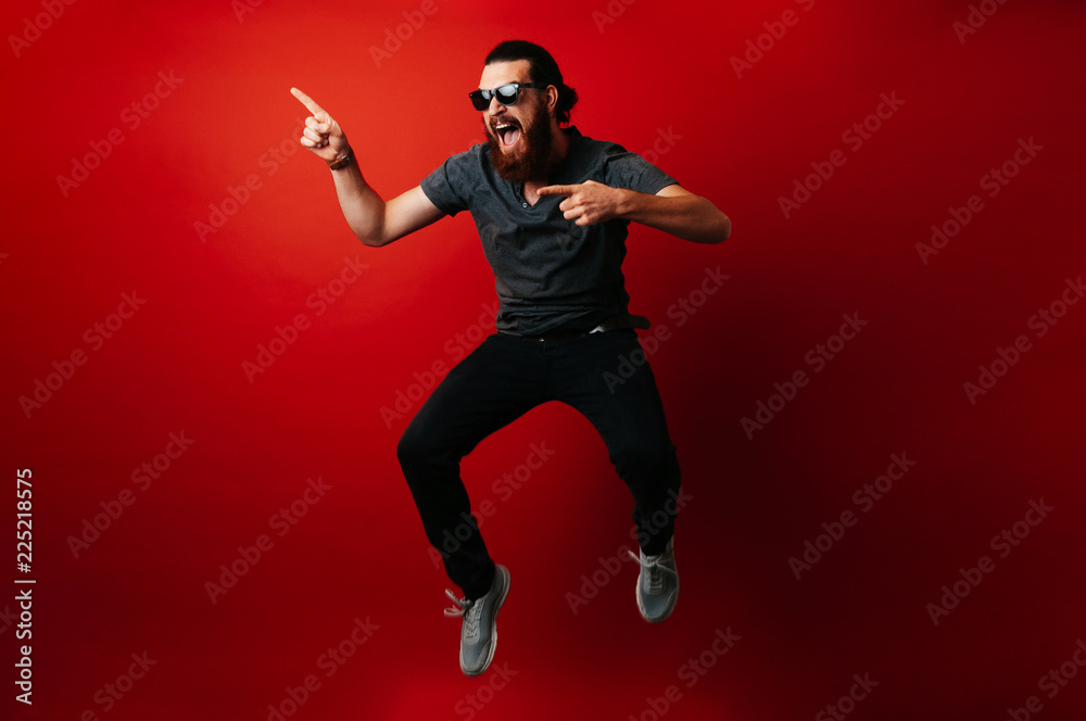 Amazed bearded hipster man wearing sunglasses and jumping over red background and pointing away