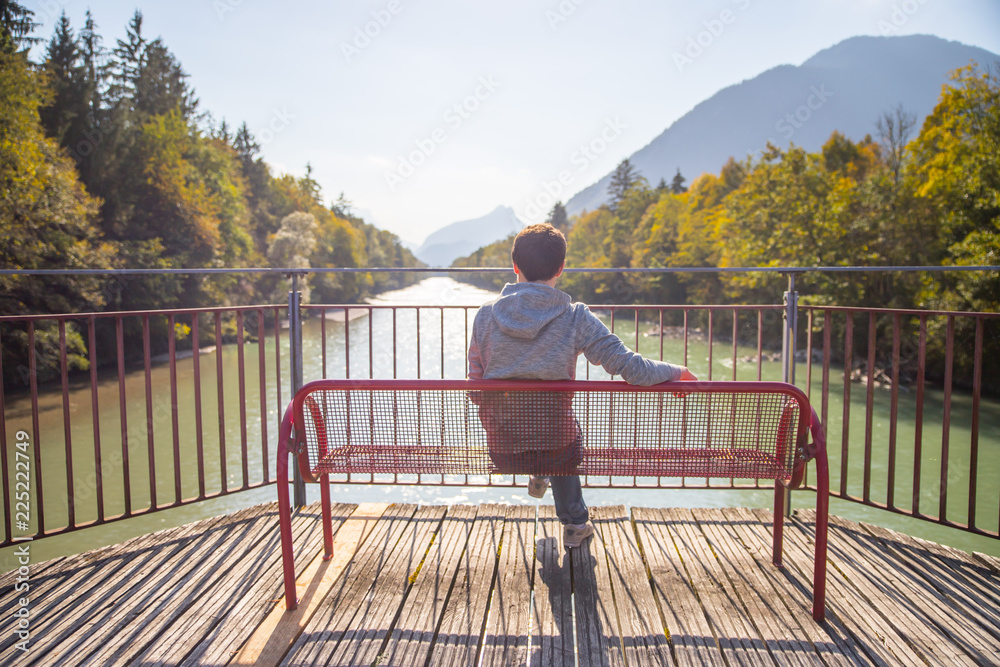 Junger Mann sitzt entspannt auf Spazierbank, Blick auf Fluss und Wälder Junger Mann sitzt entspannt auf Spazierbank, Blick auf Fluss und Wälder