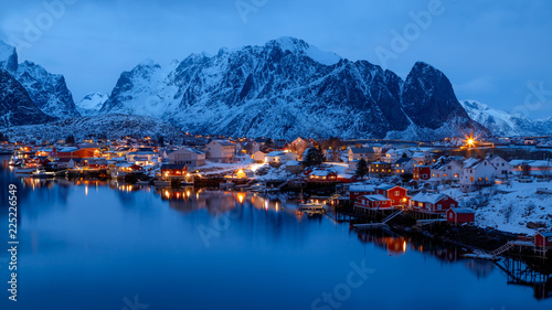 Lofoten islands  Norway. Colorful winter landscape in blue hours. Illuminated fishing village reflected in water. Snowy mountains in background. 