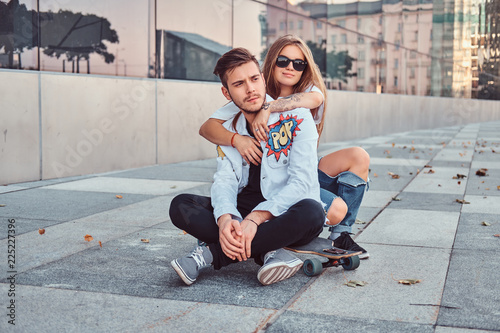 Trendy dressed young couple - pretty girl embrace her boyfriend while they sitting together on a skateboard near skyscraper.