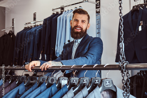Smiling tattoed male with stylish beard and hair dressed in elegant suit standing in a menswear store.