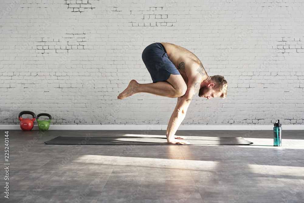 Fototapeta premium Muscular tattooed athlete exercising yoga asana push up inside in sunny sport club. Fit shirtless male fitness model..