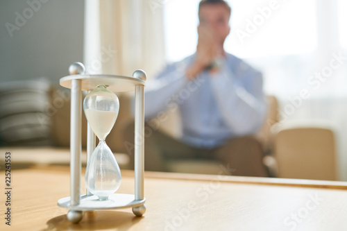 Closeup of hourglass on table with blurred shape of man waiting in background, copy space