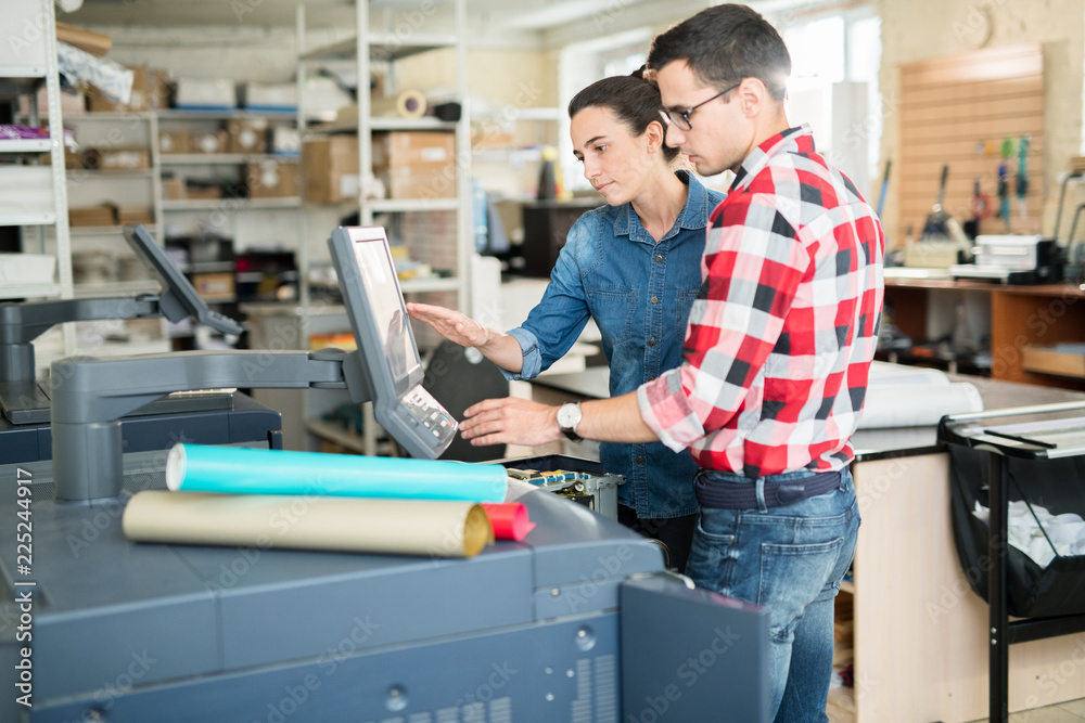 Confident experts working in printing department of publishing company, they discussing print settings while using touchscreen of printer in office Confident experts working in printing department of publishing company, they discussing print settings while using touchscreen of printer in office