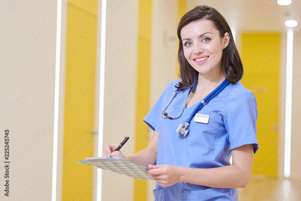 Waist up portrait of smiling young nurse looking at camera while posing in clinic holding clipboard, copy space Waist up portrait of smiling young nurse looking at camera while posing in clinic holding clipboard, copy space