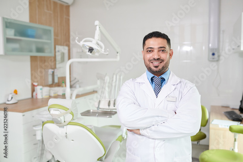 Waist up portrait of Middle-Eastern dentist posing in office smiling at camera while standing with arms crossed by dental chair  copy space