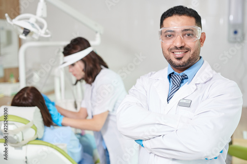 Waist up portrait of confident Middle-Eastern doctor wearing lab coat posing in dentists office with patient in background, copy space