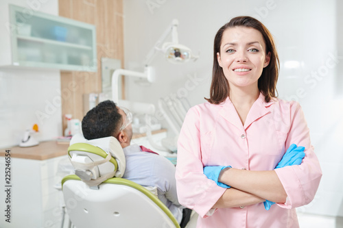 Waist up portrait of pretty female dentist looking at camera and smiling while posing standing with arms crossed in office  patient in background  copy space