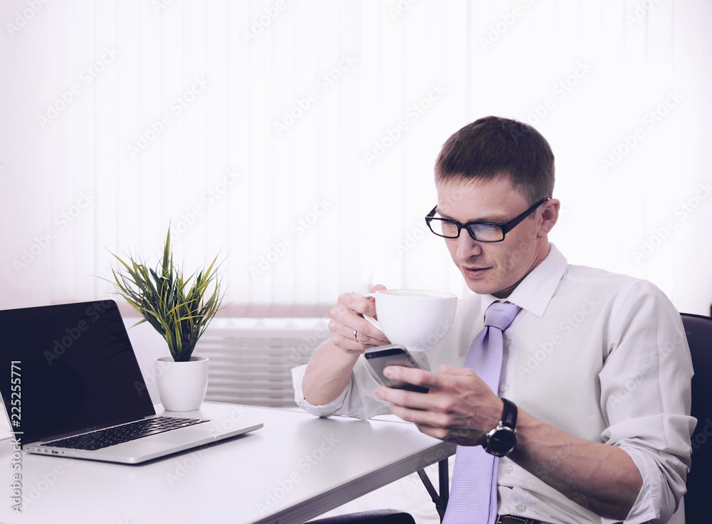 Φωτογραφία Young businessman trader sits at a table in the morning in a room near a laptop with a Cup of coffee