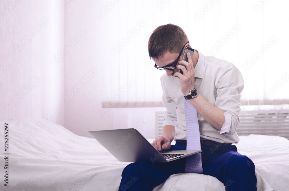 Φωτογραφία Young businessman trader sits in the morning on a white bed in a room near a laptop with a phone in his hand