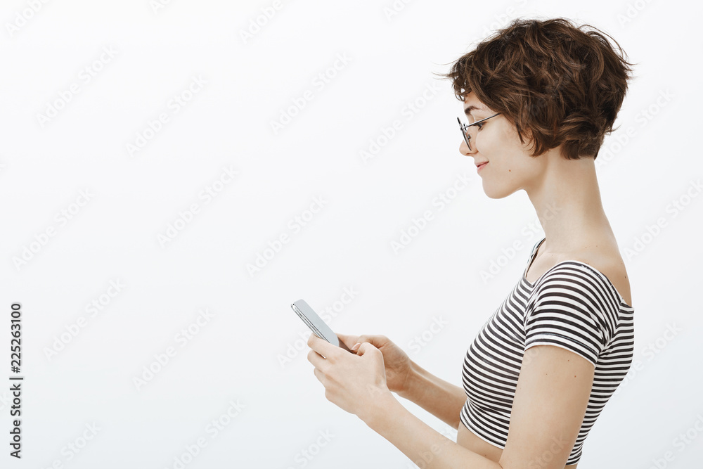 Fotka „Smart and attractive girl reading while standing in subway. Cute  stylish urban woman standing in profile, smiling while looking at  smartphone screen, playing games or messaging via social network“ ze služby