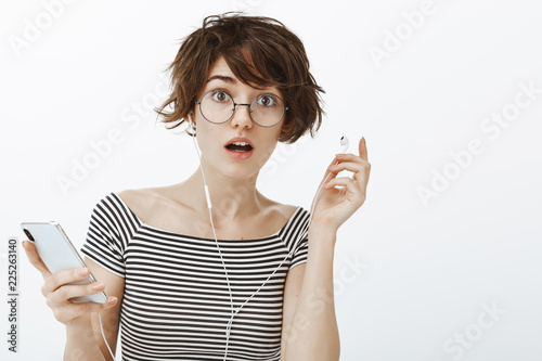 Repeat please I was in earphones. Portrait of questioned cute european woman in striped t-shirt, taking off earbud to hear question, holding smartphone, interrupted from listening music