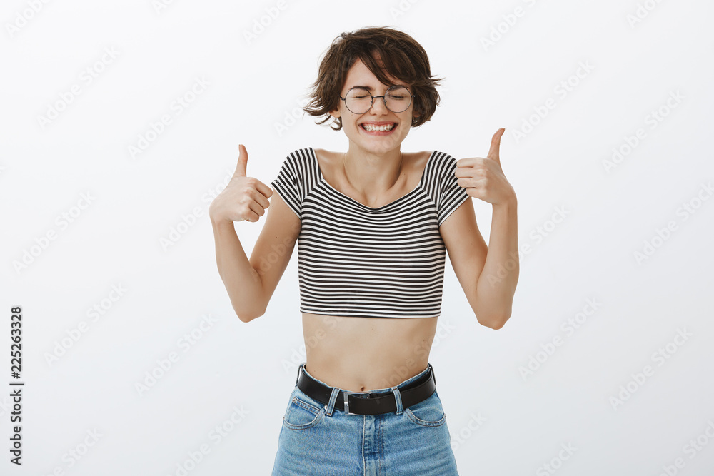 Studio shot of excited happy and pleased cute female with short brown hair, closing eyes and smiling joyfully while showing thumbs up and giving positive answer, agreeing or liking idea Studio shot of excited happy and pleased cute female with short brown hair, closing eyes and smiling joyfully while showing thumbs up and giving positive answer, agreeing or liking idea