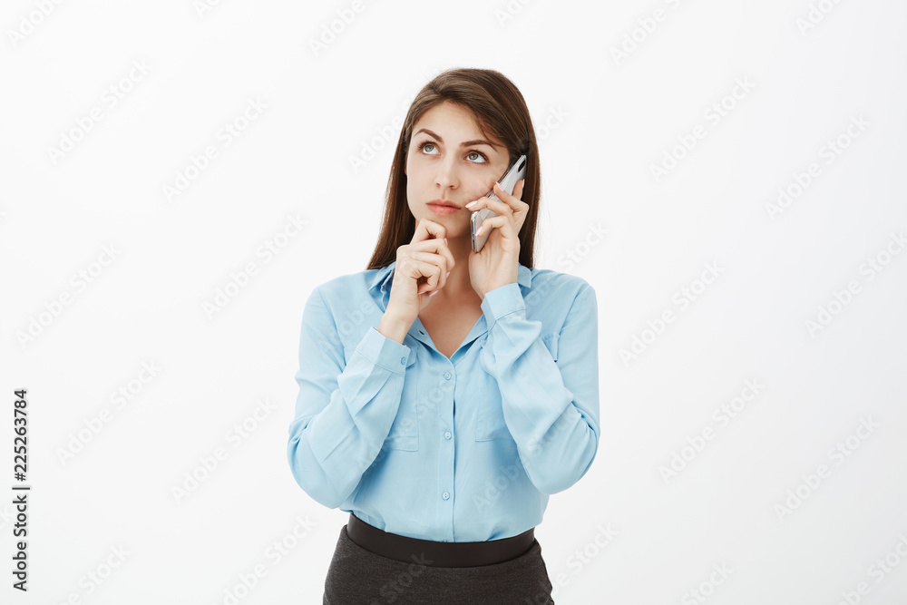 Let me think and weigh everything. Portrait of focused good-looking brunette in blue blouse, holding smartphone near ear, touching chin and looking at upper left corner, thinking, making decision Let me think and weigh everything. Portrait of focused good-looking brunette in blue blouse, holding smartphone near ear, touching chin and looking at upper left corner, thinking, making decision