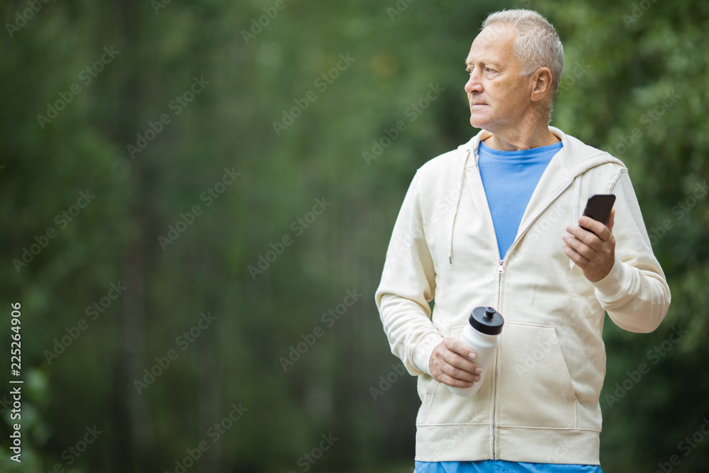 Fototapeta Starszy aktywny mężczyzna w odzieży sportowej, trzymając smartfon i butelkę wody podczas porannego treningu w parku