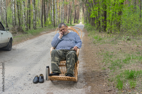 Tired senior driver man resting outside of his car sleeping in a wicker rocking-chair in spring forest