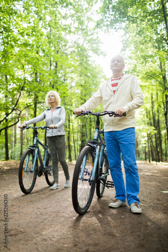 Retired couple in activewear standing on road in park by their bicycles
