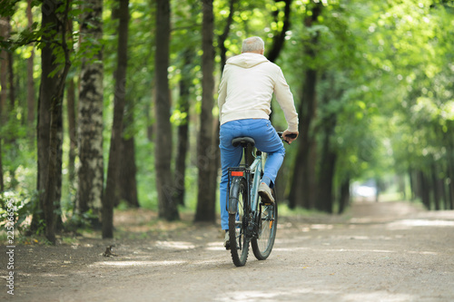 Rear view of active senior man riding bicycle along forest road on summer day