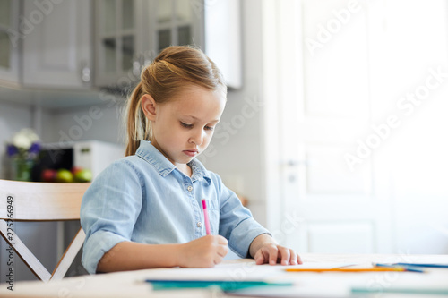 Little girl concentrating on drawing with crayons while sitting in the kitchen at home