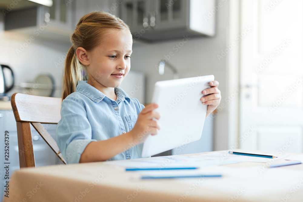 Happy little girl watching videos on touchpad screen while sitting by table at home Happy little girl watching videos on touchpad screen while sitting by table at home