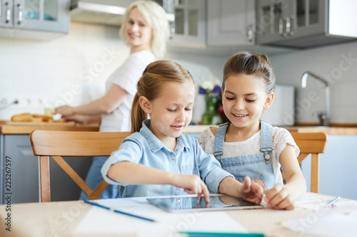 Two sisters in casualwear sitting by table and playing game in tablet while their mom cooking dinner on background