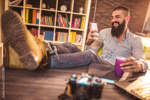 Portrait of modern young man relaxing in office with feet on desk texting via smartphone photo