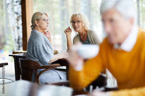 Content attractive mature ladies in casual clothing sitting at table and gossiping about handsome man in cafe