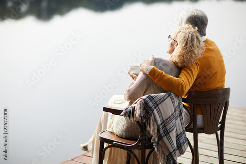 Rear view of dreamy fashionable senior couple sitting on chairs and relaxing with coffee cup on pier, elderly man embracing beloved woman at romantic date