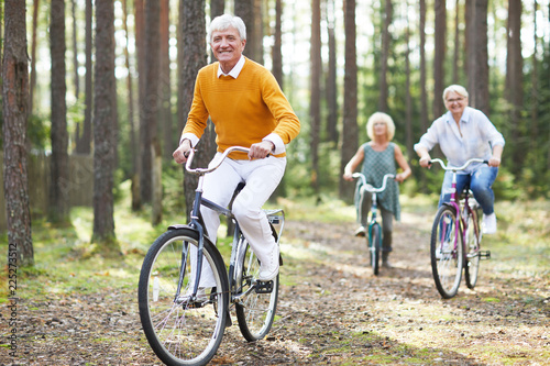Cheerful excited senior friends in casual clothing enjoying active life cycling on bikes together in forest