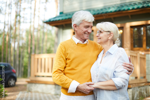 Positive romantic senior couple embracing and holding hands while looking into eyes, they standing against cottage