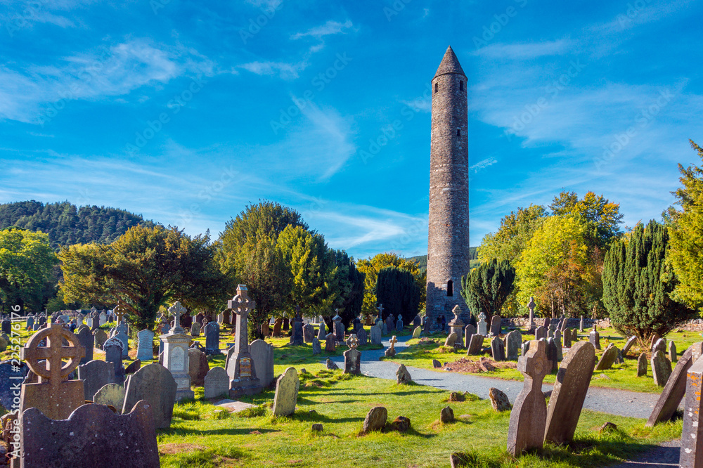 Naklejka premium Kamienna okrągła wieża i niektóre ruiny osady klasztornej pierwotnie zbudowanej w VI wieku w dolinie Glendalough, hrabstwo Wicklow, Irlandia