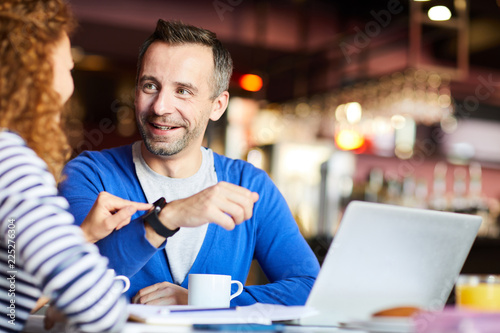 Cheerful confident handsome man with stubble showing new smartwatch to colleague who touching screen and viewing function of device in cafe
