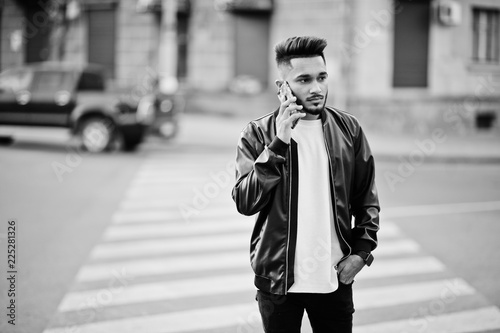 Stylish indian beard man at black leather jacket walking on zebra crossing. India model posed outdoor at streets of city and speaking phone.