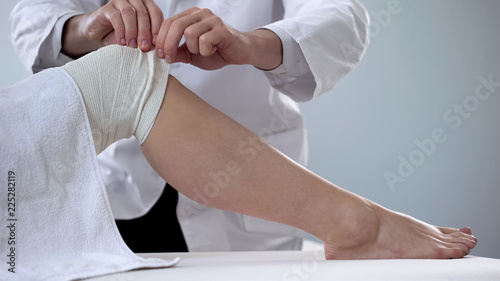 Traumatologist bandaging patients sprained knee, first aid for injuries, clinic