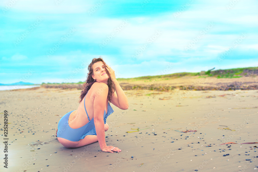 Young brunette girl in striped swimsuit sitting on the beach on the sand Shining bright sun blue sky clouds