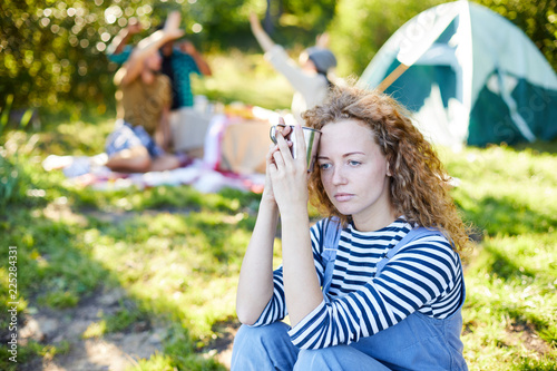 Young serene woman with mug sitting on ground and relaxing while her friends having fun on background