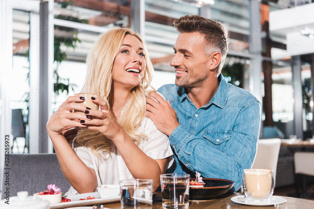 boyfriend hugging laughing girlfriend and she holding cup of coffee at table in cafe