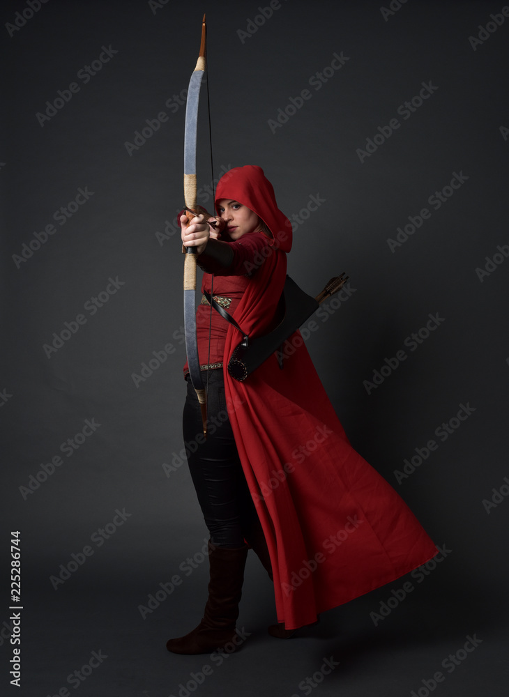 full length portrait of brunette girl wearing red medieval costume and cloak, holding a bow and arrow. standing pose on grey studio background.