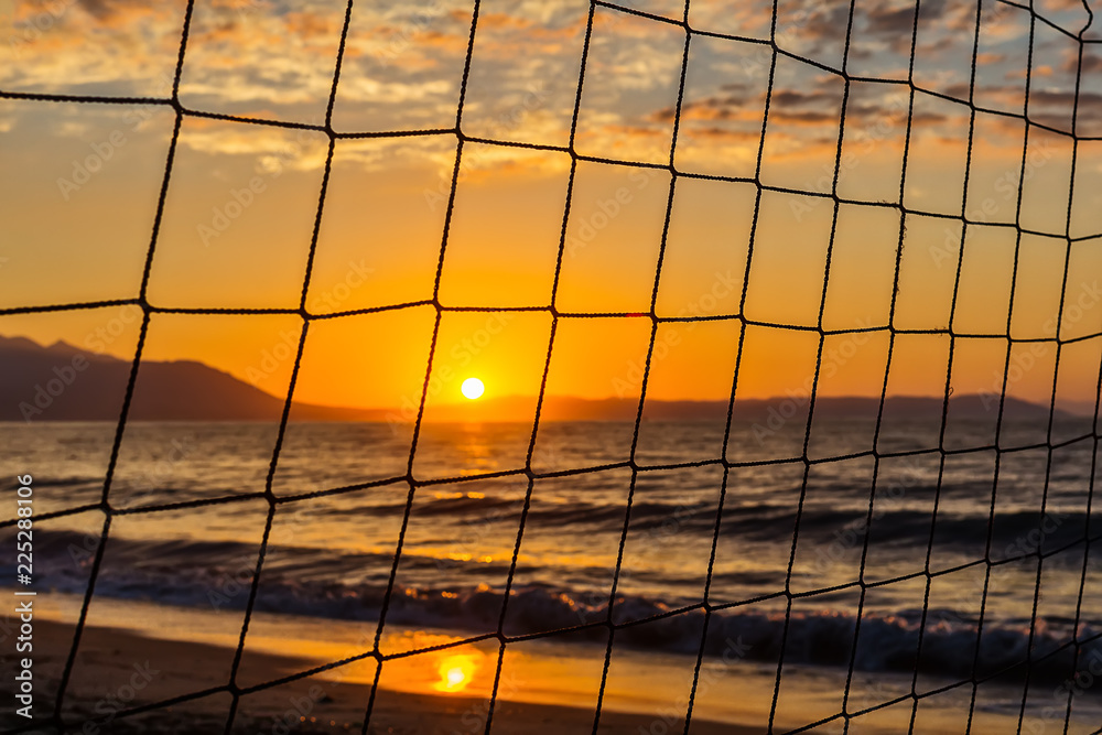 Naklejka premium View of the sunrise through the volleyball net. Early morning , dramatic sunrise over sea and mountain.