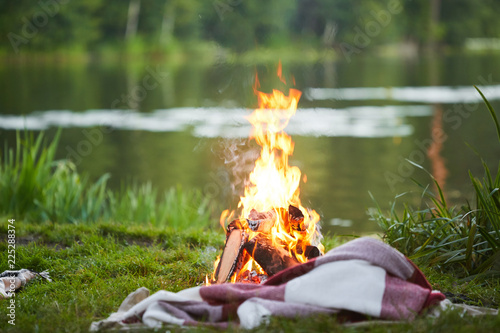 Burning campfire and plaid on green grass by river or lake in natural environment