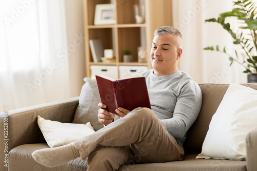 leisure, literature and people concept - man sitting on sofa and reading book at home