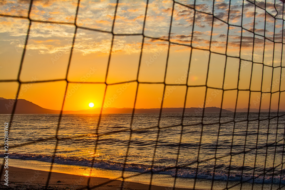 Naklejka premium View of the sunrise through the volleyball net. Early morning , dramatic sunrise over sea and mountain.