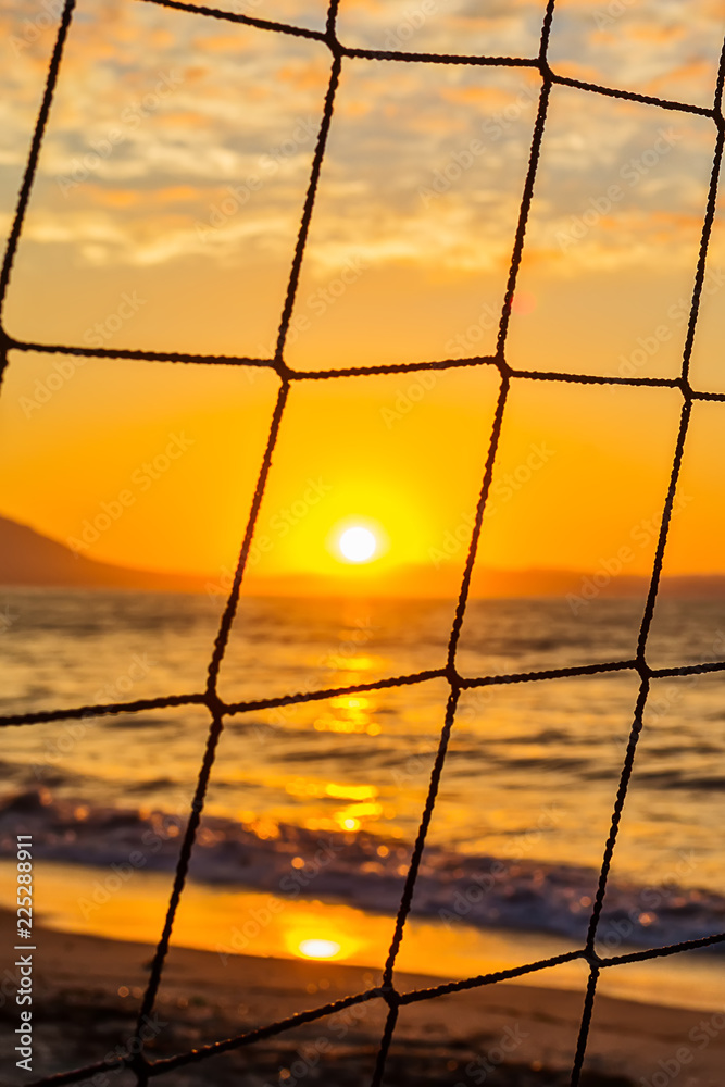 Fototapeta premium View of the sunrise through the volleyball net. Early morning , dramatic sunrise over sea and mountain.