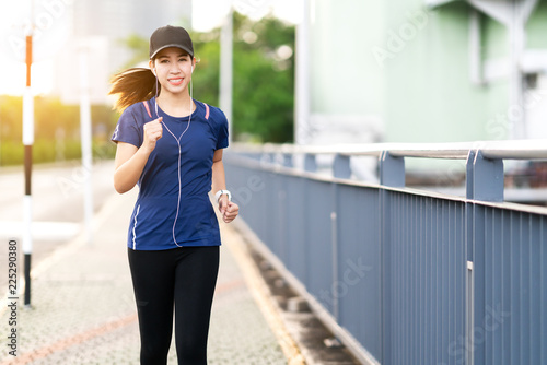 Young attractive asian runner woman running in city street or foot path way listen to music wearing blue or black sporty gadget in leisure sport concept with sun light in morning or evening workout.