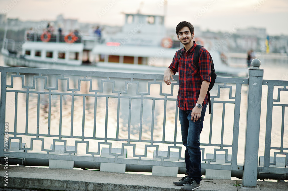 Young indian student man at checkered shirt and jeans with backpack posed on evening city against lake and boat.