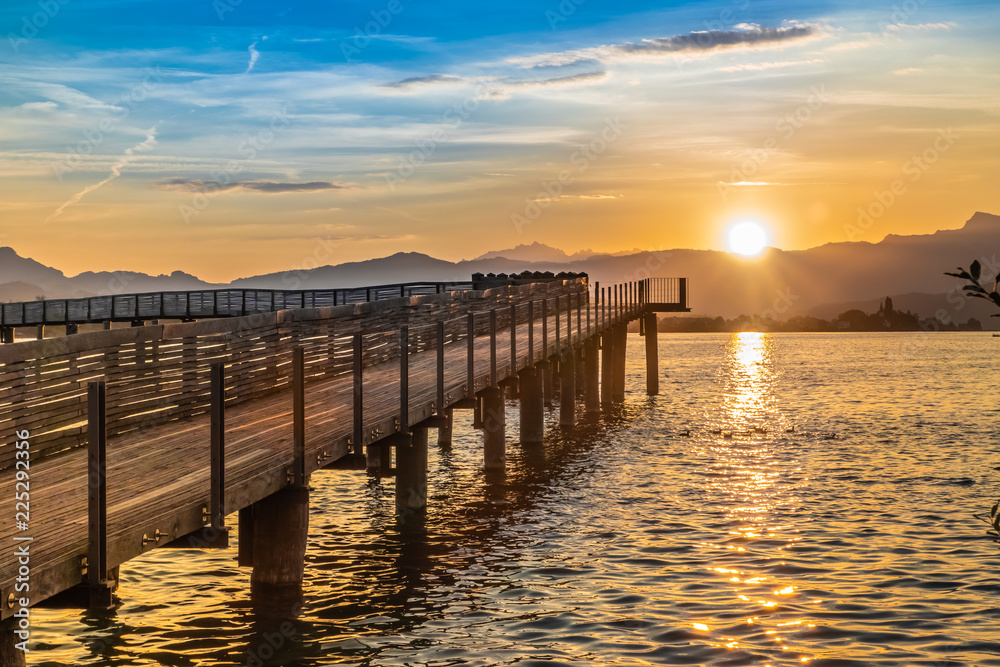 Fototapeta premium Dramatic sunrise during the Fall Equinox over the historical holzsteg pedestrian bridge crossinhg the Upper Zurich Lake (Obersee), part of the old Way of Saint James, Switzerland