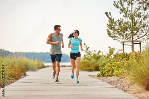 fitness, sport and lifestyle concept - happy couple in sports clothes and sunglasses running along summer beach path