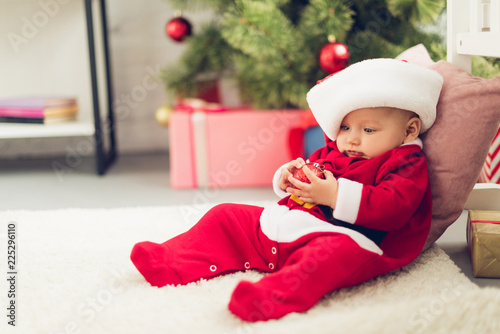 adorable little baby in santa suit sitting on floor with blurred christmas tree on background