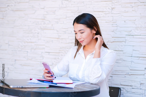 Asian bisiness woman dialing number on cellphone using good internet connection indoors. Kazakh female puzzled on watching video in social networks via cellular while sitting in cafe. photo