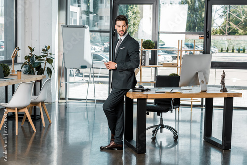 smiling handsome businessman leaning on table and holding smartphone in office